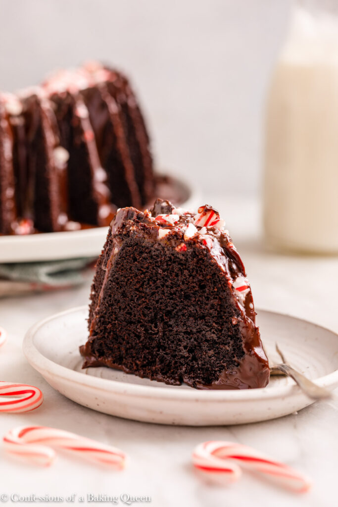 slice of peppermint chocolate cake on a white plate with a fork with cake and milk in the background on a white marble surface with candy canes
