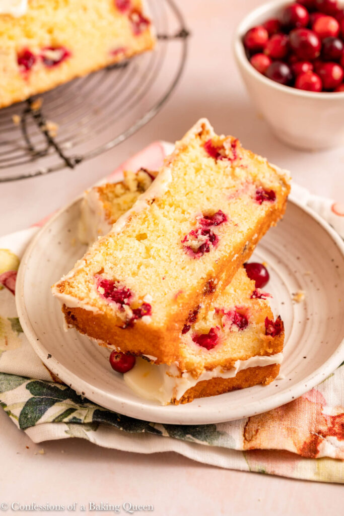 two slices of cranberry orange loaf cake on a white plate on top of a floral linen on a light pink surface
