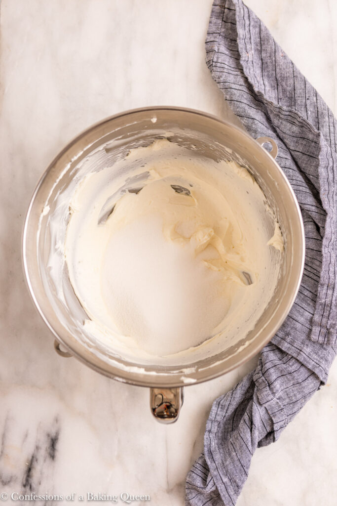 whipped cream cheese with sugar added in a metal mixing bowl on a white marble surface with a blue linen