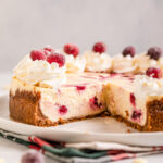 cut open white chocolate cranberry cheesecake on a white plate on top of a christmas linen on a light grey surface