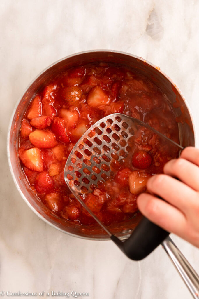 potato masher lightly mashing some strawberries in a pot on a white marble surface