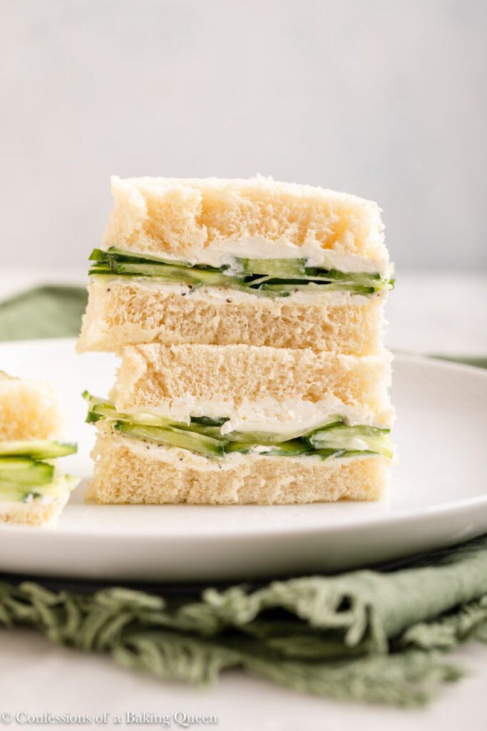 afternoon tea sandwiches on a white plate, two cucumber and cream cheese sandwiches stacked on a white plate on a white marble surface with a green linen