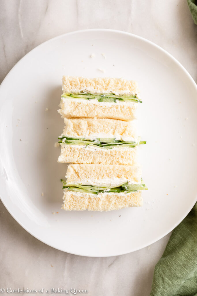 afternoon tea sized cucumber and cream cheese sandwiches on a white plate on a white marble surface with a green linen