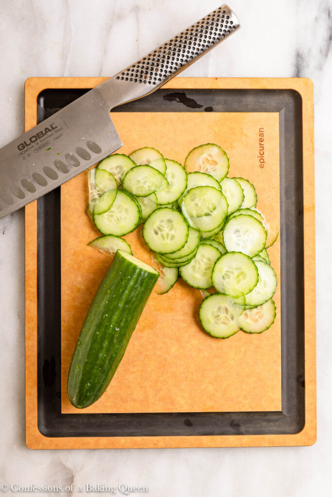 knife cutting cucumbers into thin slices on a wood cutting board on a white marble surface