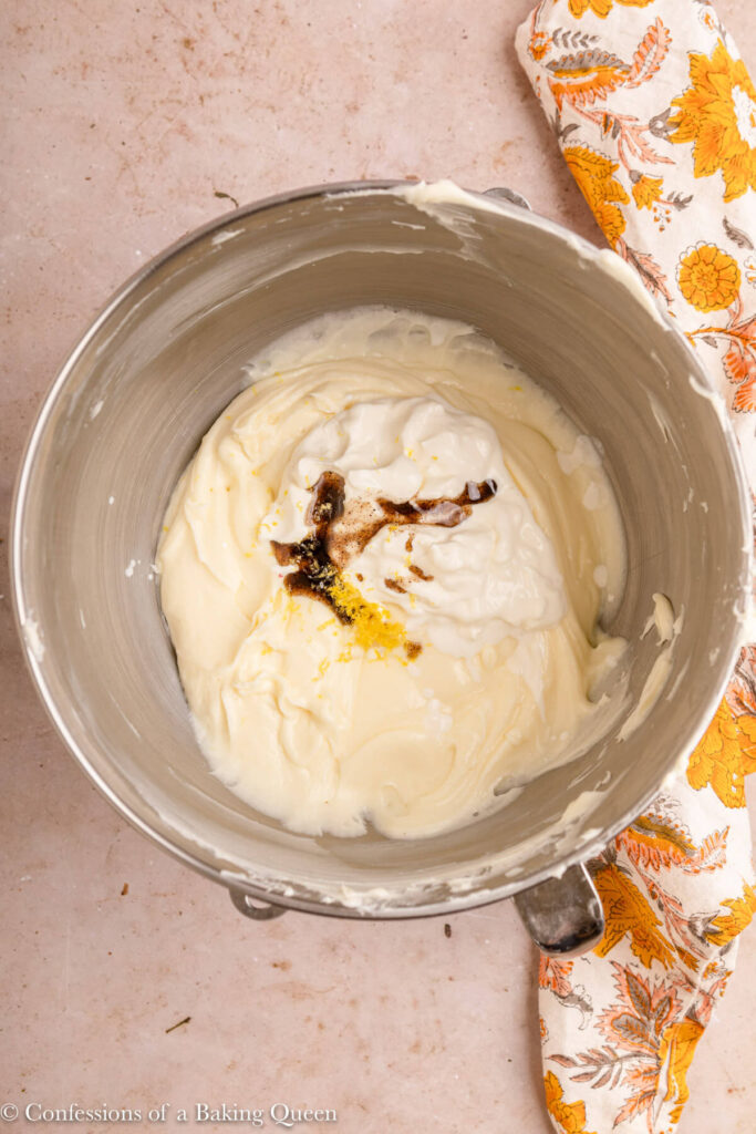 sour cream, vanilla bean paste and lemon zest added to cream cheese mascarpone mixture in a metal mixing bowl on a light brown surface with a yellow floral linen