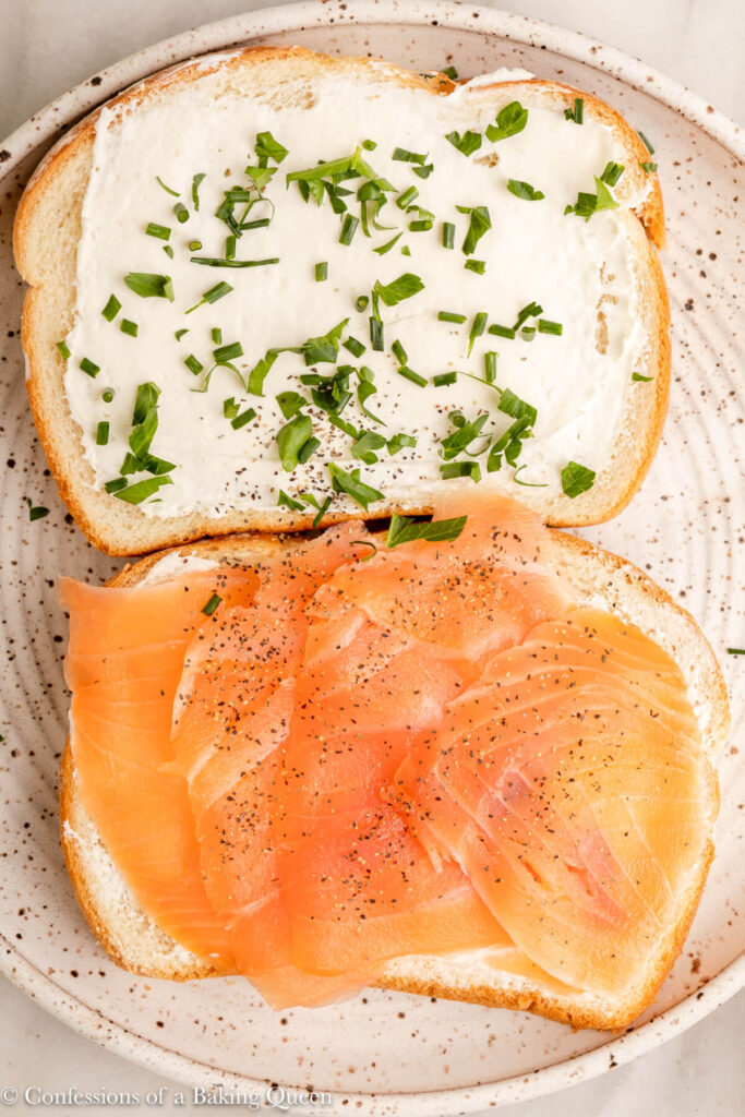 close up of cream cheese spread on bread with fresh herbs and smoked salmon on a white plate on a marble surface