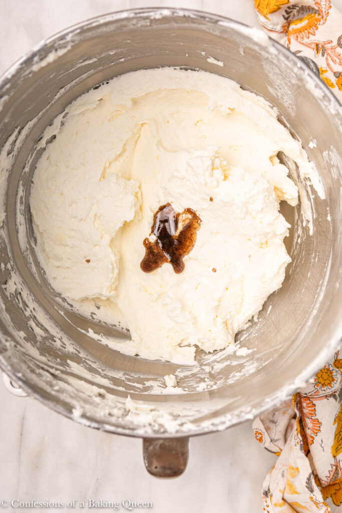 vanilla bean paste added to italian merinuge buttercream in a metal bowl on a white marble surface with a floral linen