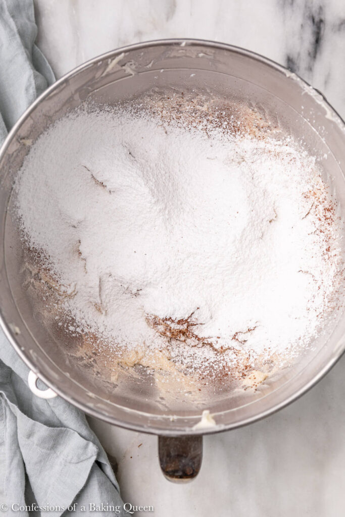 cocoa powder and confectioners sugar sifted into frosting mixture in an metal bowl on a white marble surface with a light blue linen