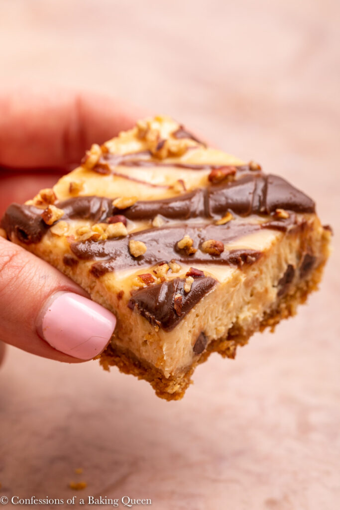 hand holding a turtle cheesecake bar up to the camera on a light brown surface