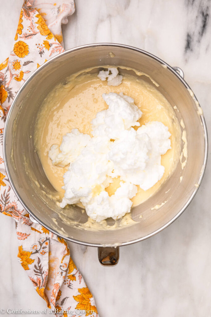 whipped egg whites added to yellow cake batter in a metal mixing bowl on a white marble surface with a yellow flowery linen