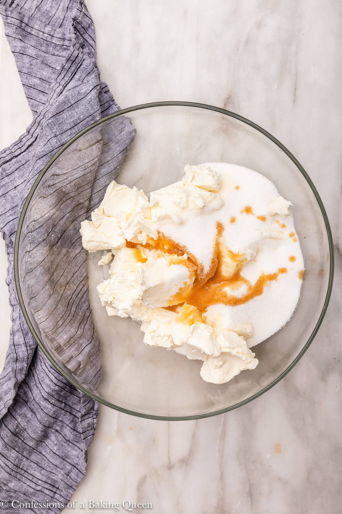 cream cheese, sugar, and vanilla mixed together in a glass bowl on a white marble surface with a blue linen