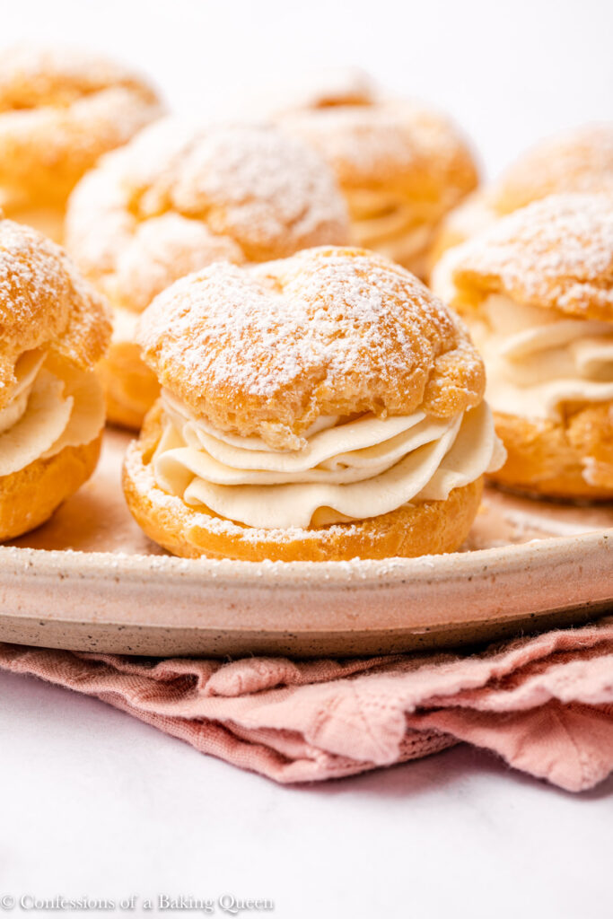 lots of cream puffs served on a light pink plate on top of a pink linen on a white surface