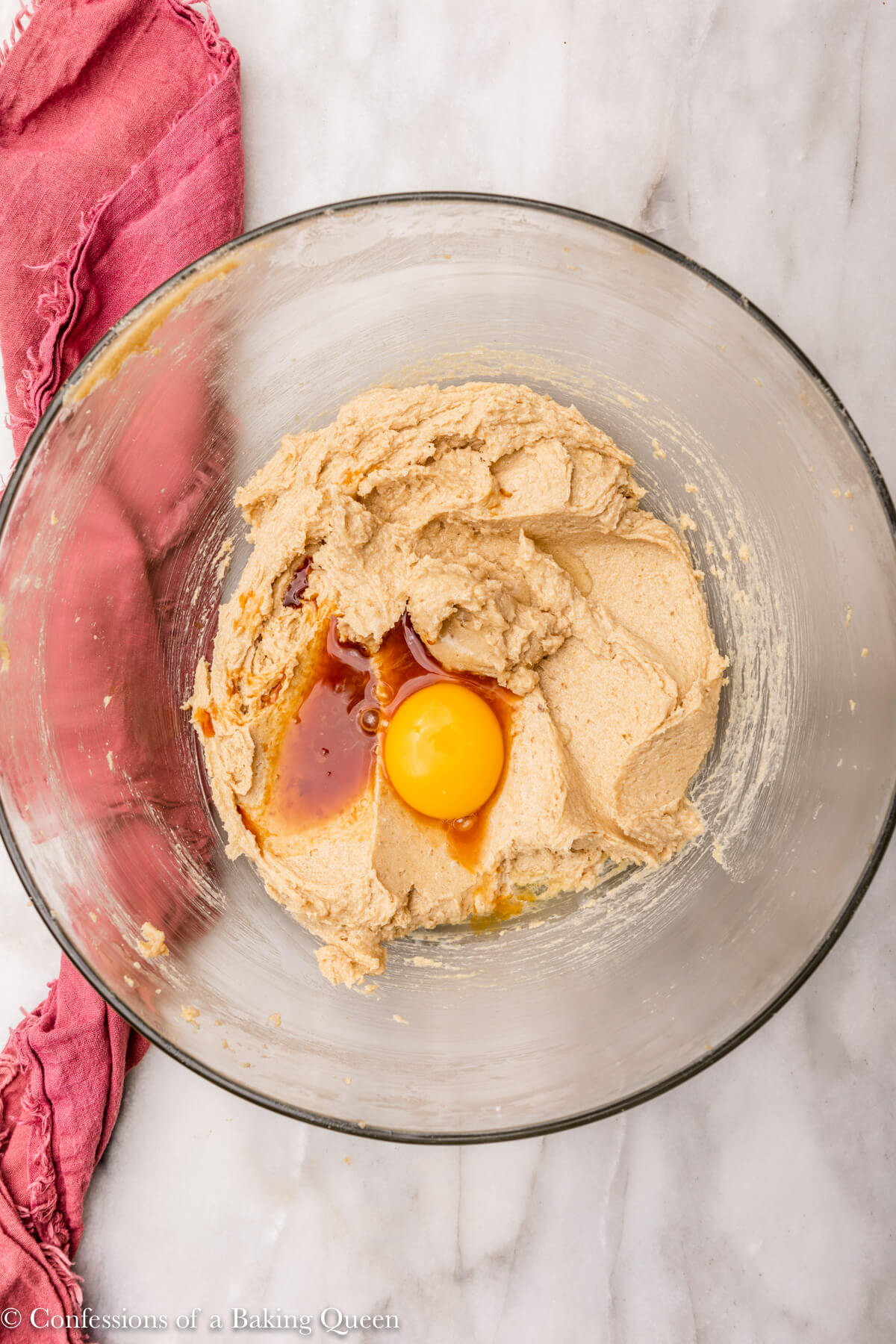 egg yolk and vanilla extract added to wet ingredients for cookies in a glass bowl next to a pink linen on a white marble surface