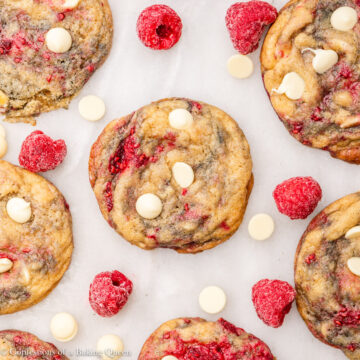 white chocolate raspberry cookies with frozen raspberries and white chocolate chips next to the cookies on a white marble surface