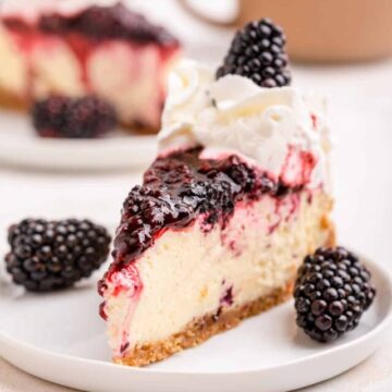 slice of blackberry cheesecake on a white plate with berries on an light surface with a brown coffee cup