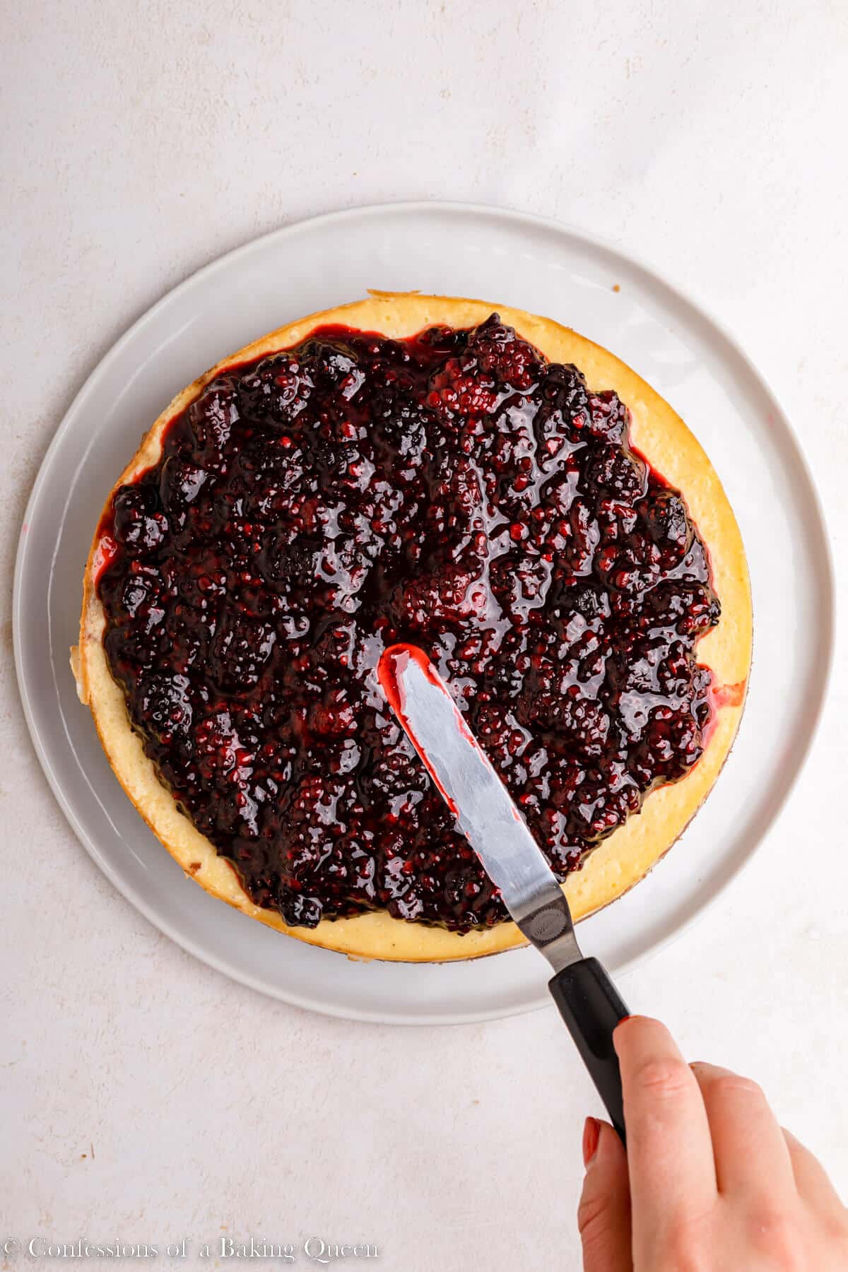 spatula spreading blackberry compote into an even layer on cheesecake on a light surface