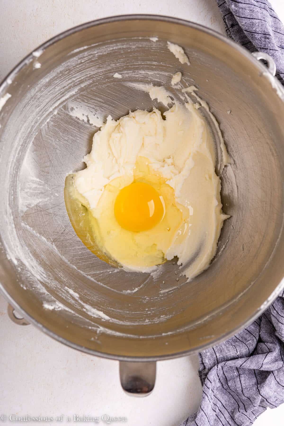 egg added to creamed sugar and butter in an large metal bowl on alight surface with a blue linen