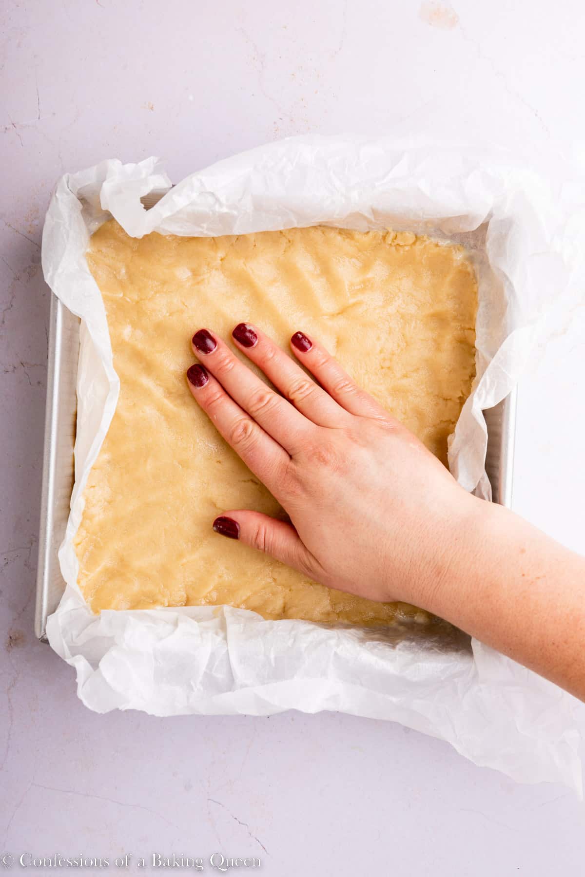 hand pressing shortbread dough into an even layer in a square baking tin on a light pink surface