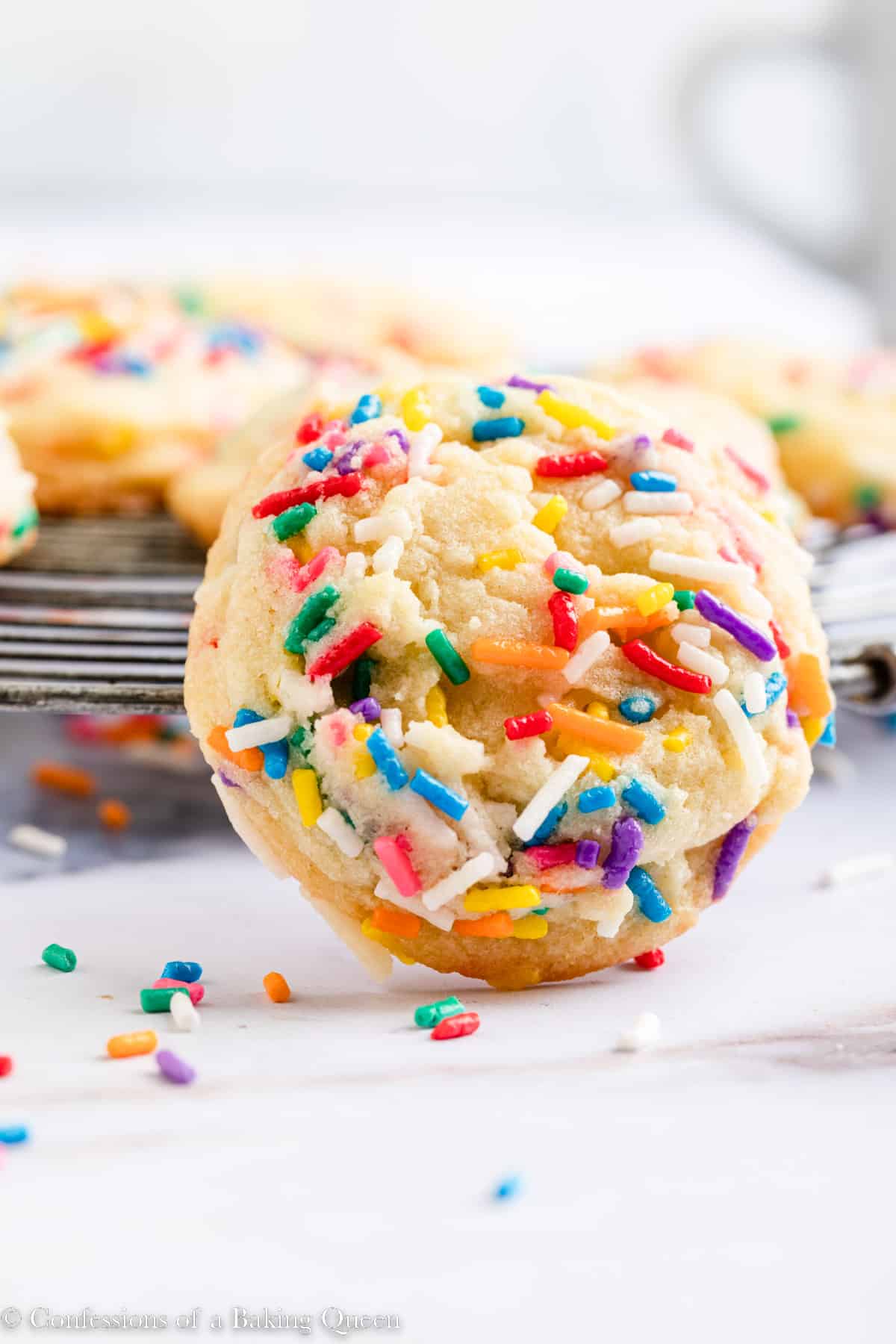 close up of funfetti cookie leaning against a wire rack on a white surface with a coffee cup in the background