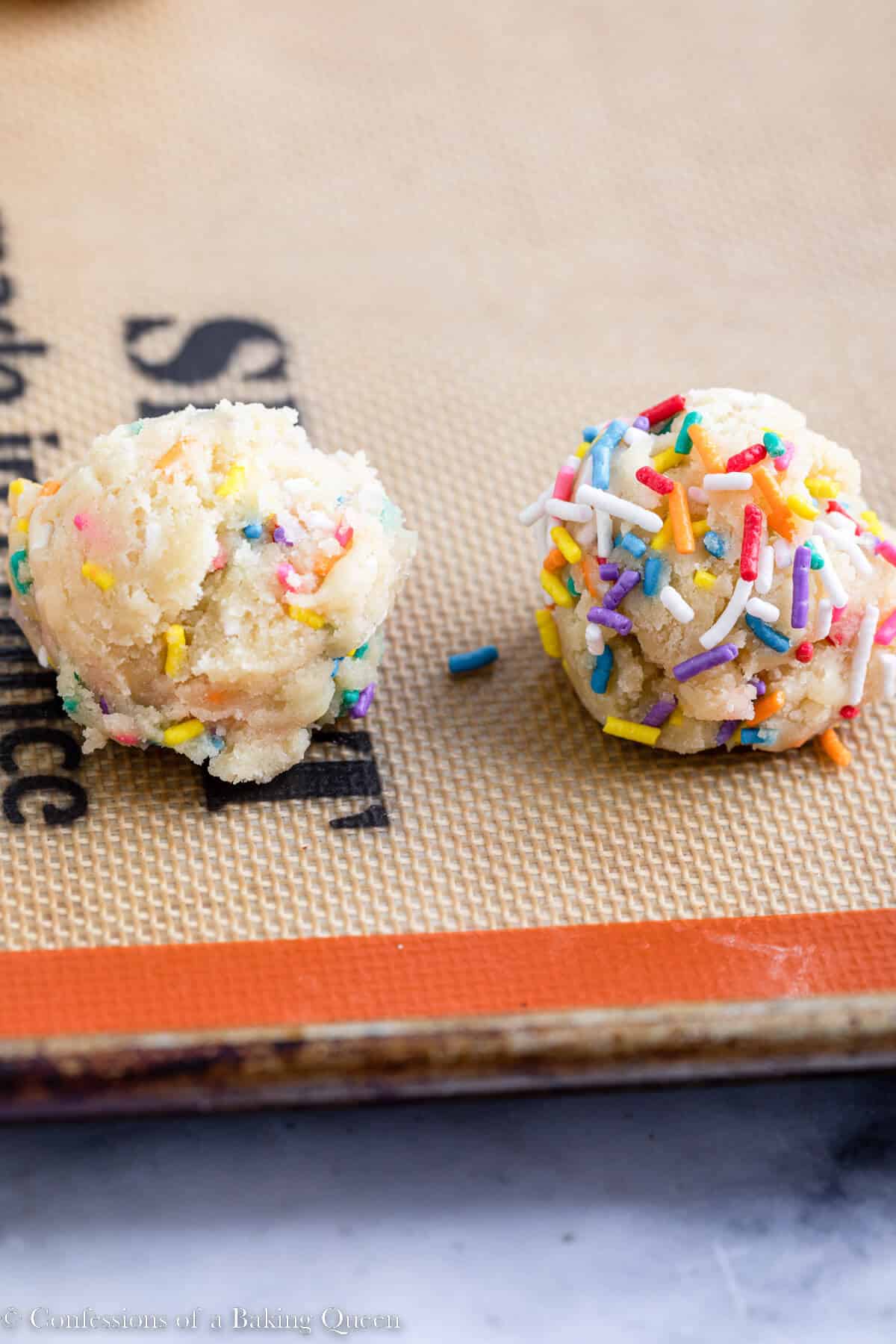 sprinkle covered cookie dough ball versus non sprinkled cookie dough ball on a silpat lined cookie sheet on a white marble surface