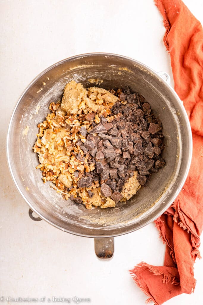 toasted walnuts and chocolate chips added to cookie dough in a metal mixing bowl on a light surface with an orange linen