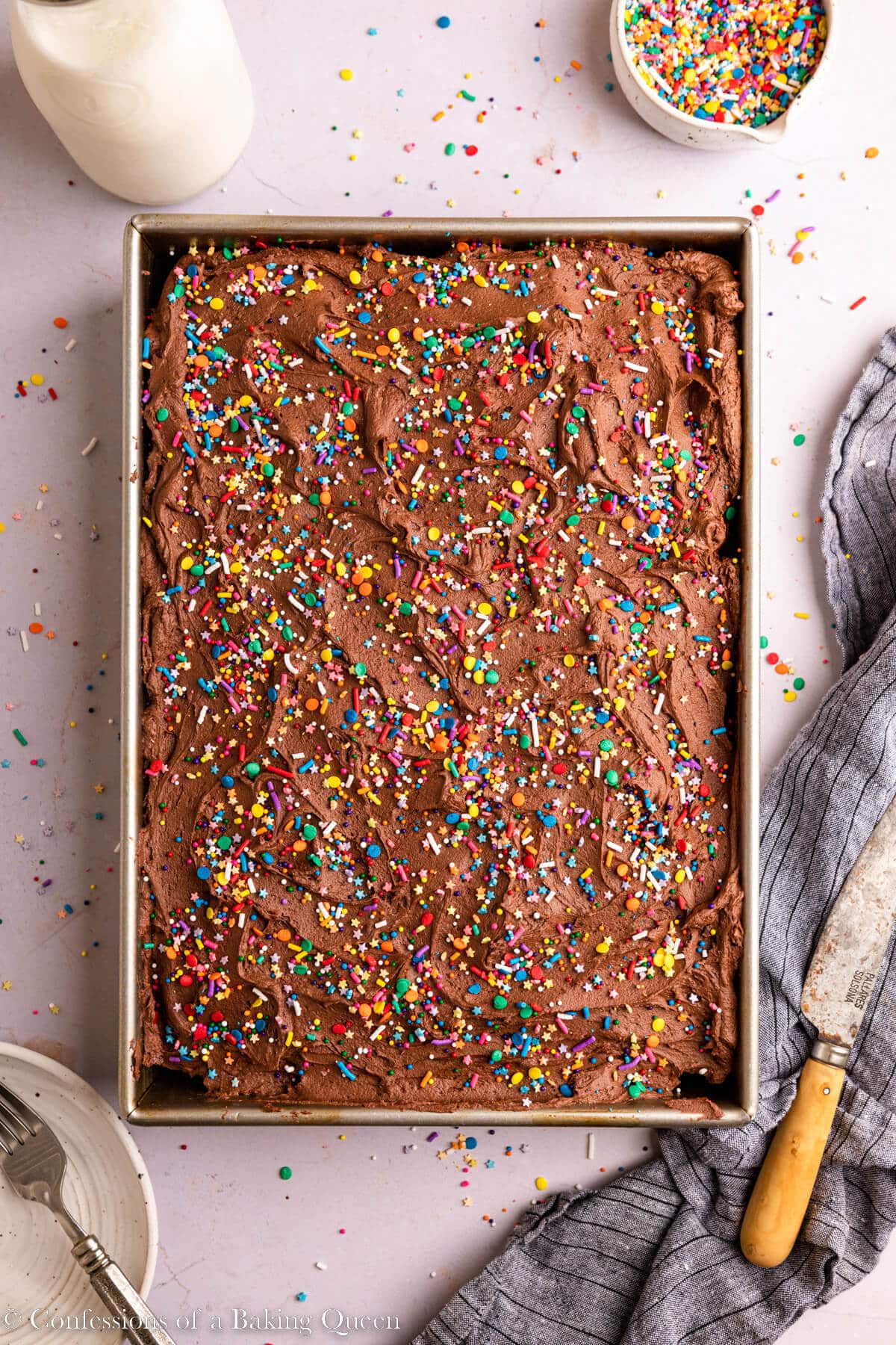 chocolate sheet cake decorated with sprinkles in a metal pan on a light surface with a blue linen, cup of sprinkles, glass of milk and plate with fork