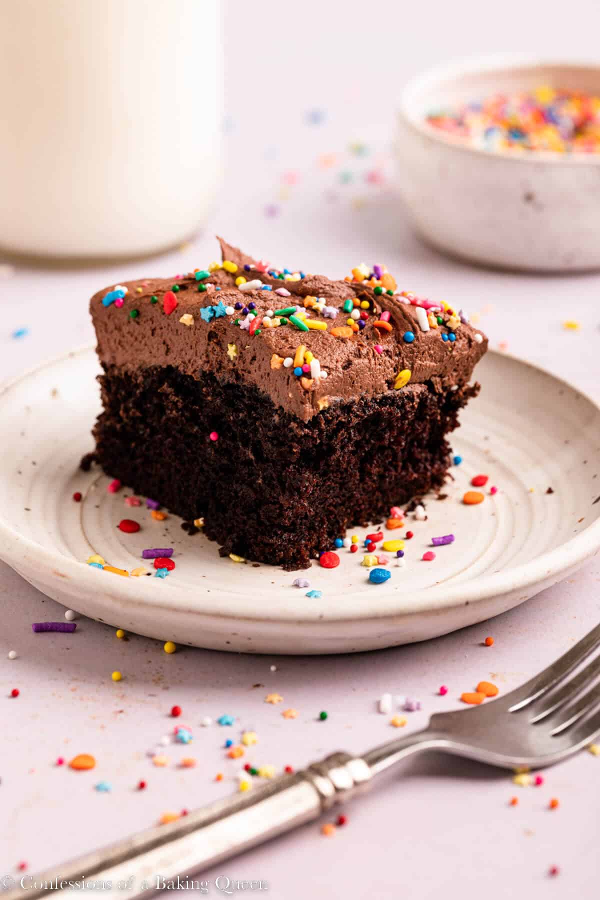 chocolate cake slice on a white plate with sprinkles on a light surface with a fork, bowl of sprinkles and glass of milk