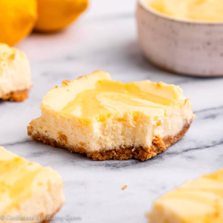 Individual slices of lemon cheesecake bars on a marble counter with fresh lemons. One slice has a bight taken out of it.