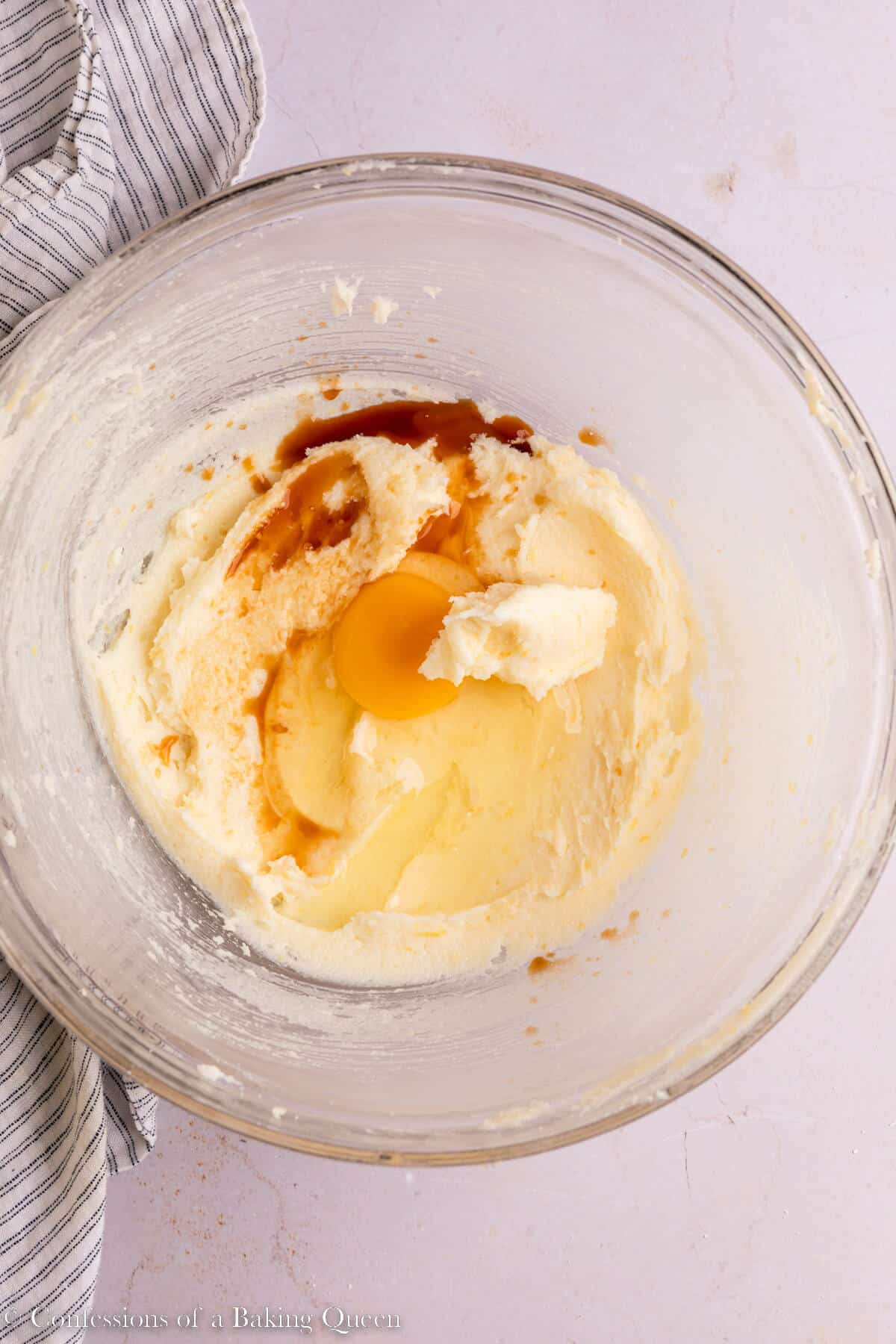 egg and vanilla extract added to creamed butter in a glass bowl on a light surface with a stripped linen