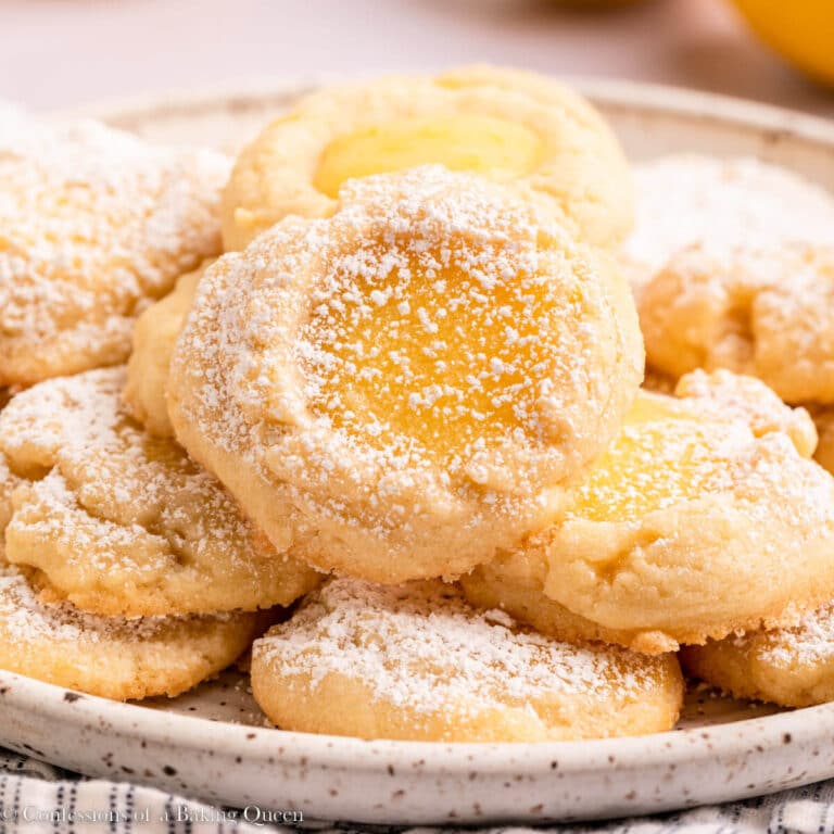 lemon curd cookies dusted in powdered sugar on a white plate on top of a stripped linen on a light surface with more lemons
