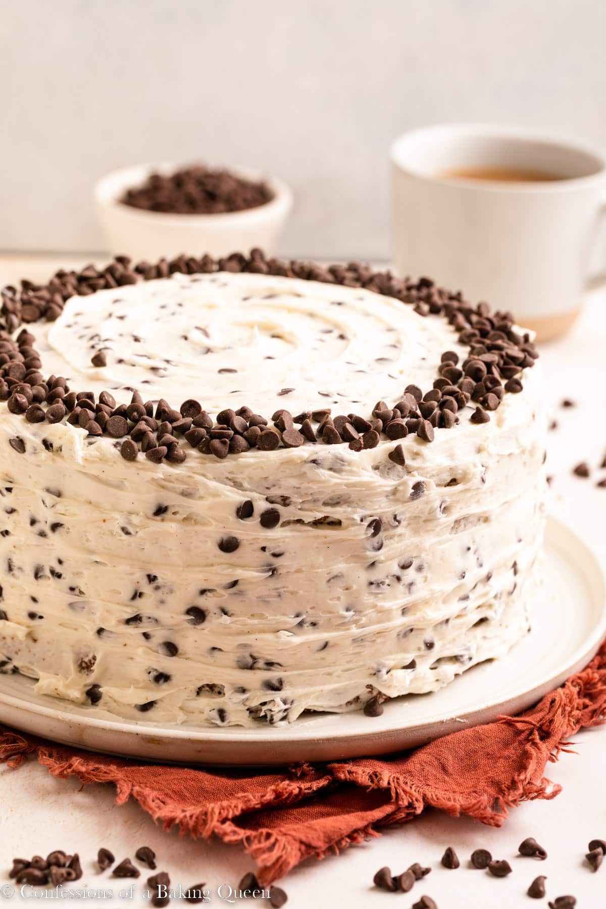 Chocolate chip cake on a serving platter near a cup of coffee and a bowl f mini chocolate chips.