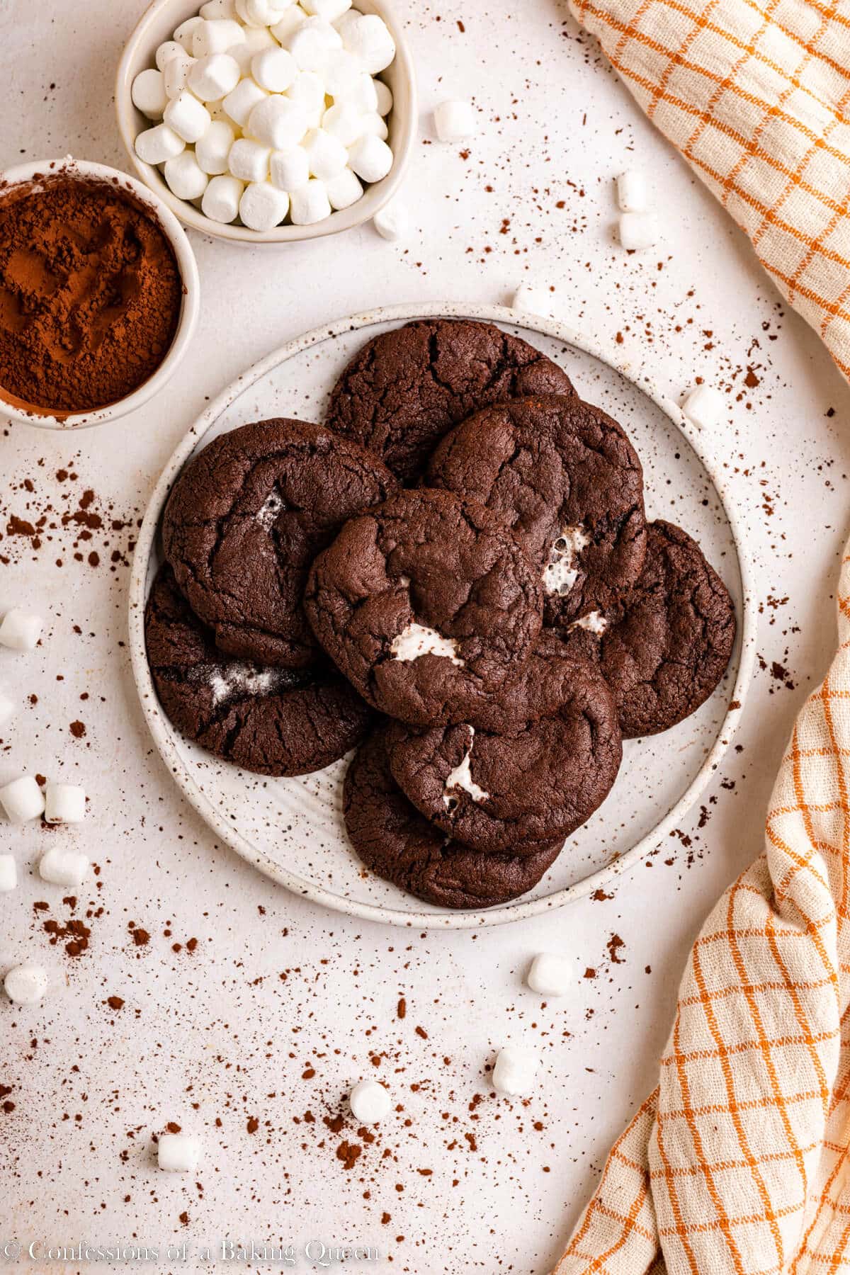 chocolate marshmallow cookies on a white plate next to a cup of marshmallows and cocoa powder on a light surface with a white and orange linen