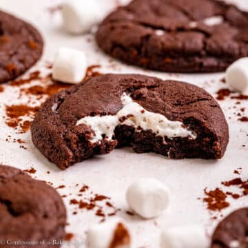 half eaten chocolate marshmallow cookie next to more cookies, marshmallows and cocoa powder on a light surface