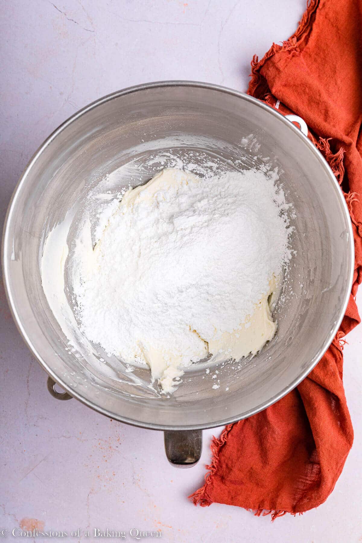 powdered sugar added to cream cheese mixture in a metal mixing bowl on a light pink surface with an orange linen