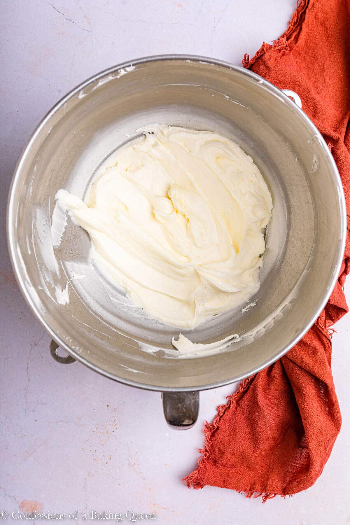 cream cheese, butter, and powdered sugar mixed together in a metal mixing bowl on a light pink surface with an orange linen