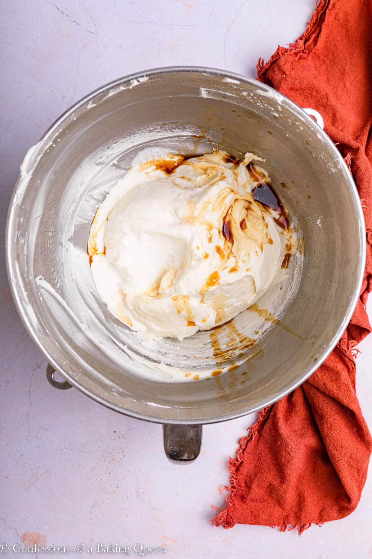 heavy cream, vanilla extract and salt added to cream cheese frosting in a metal mixing bowl on a light pink surface with an orange linen