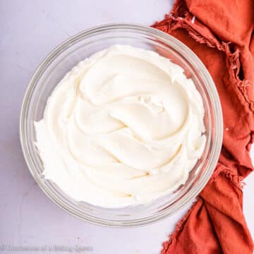 glass bowl of cream cheese frosting on a light pink surface with an orange linen