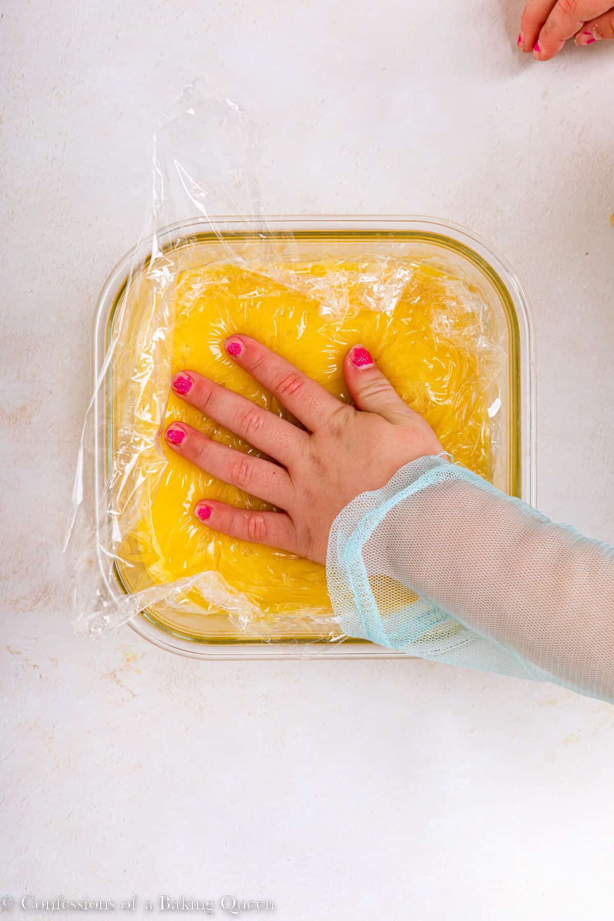 toddler hand pressing plastic wrap on to key lime curd in a glass dish