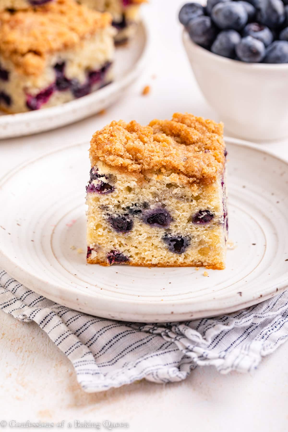 A slice of blueberry coffee cake on a plate near a bowl of fresh blueberries.