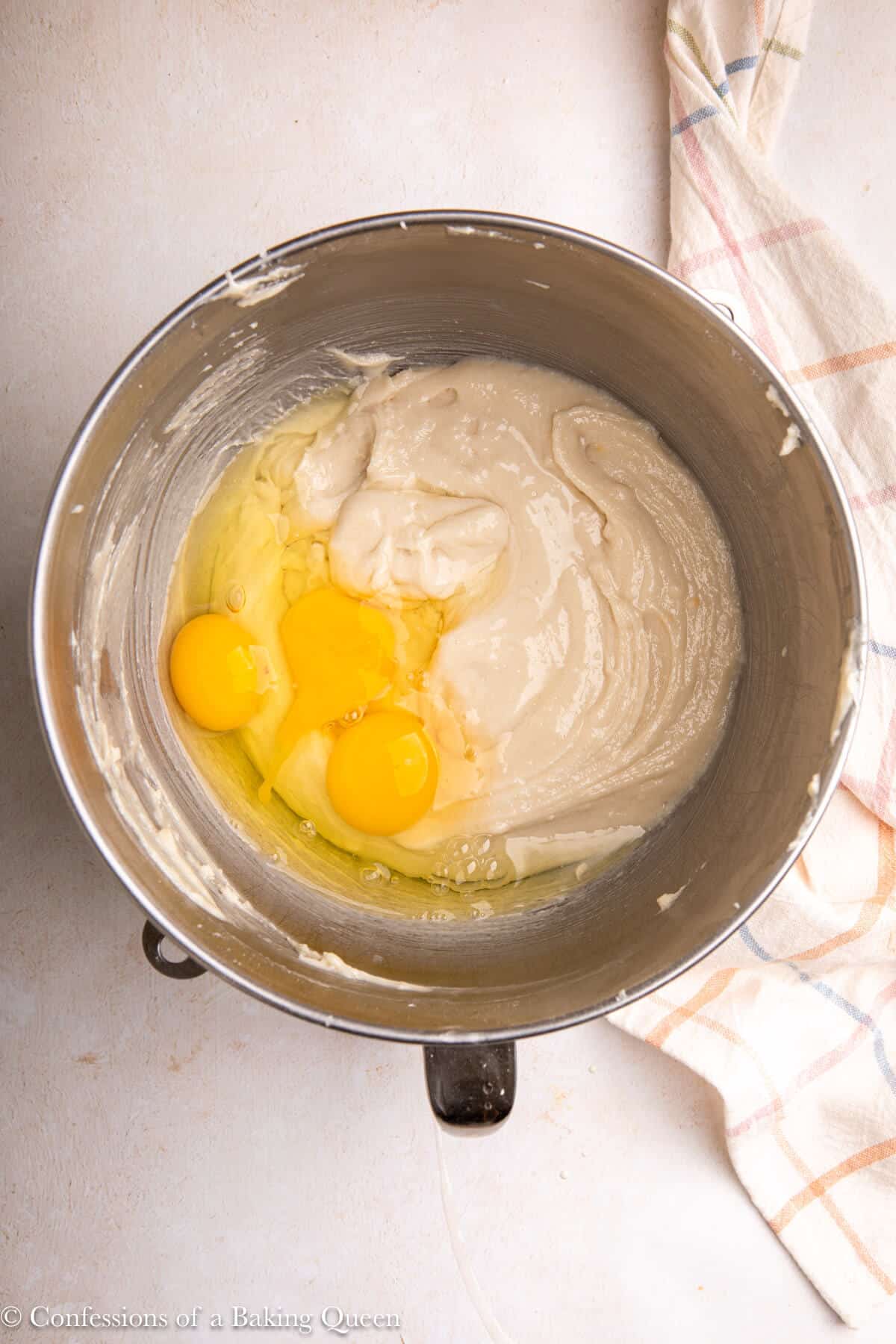 eggs added to cake batter in a metal bowl on a light surface with a checkered towel
