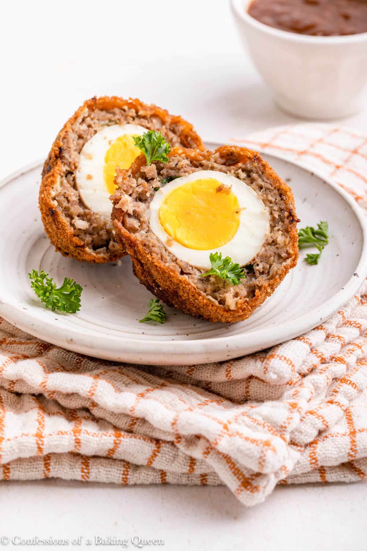 scotch egg cut into two pieces on a light white plate on top of a white and orange linen on a light surface