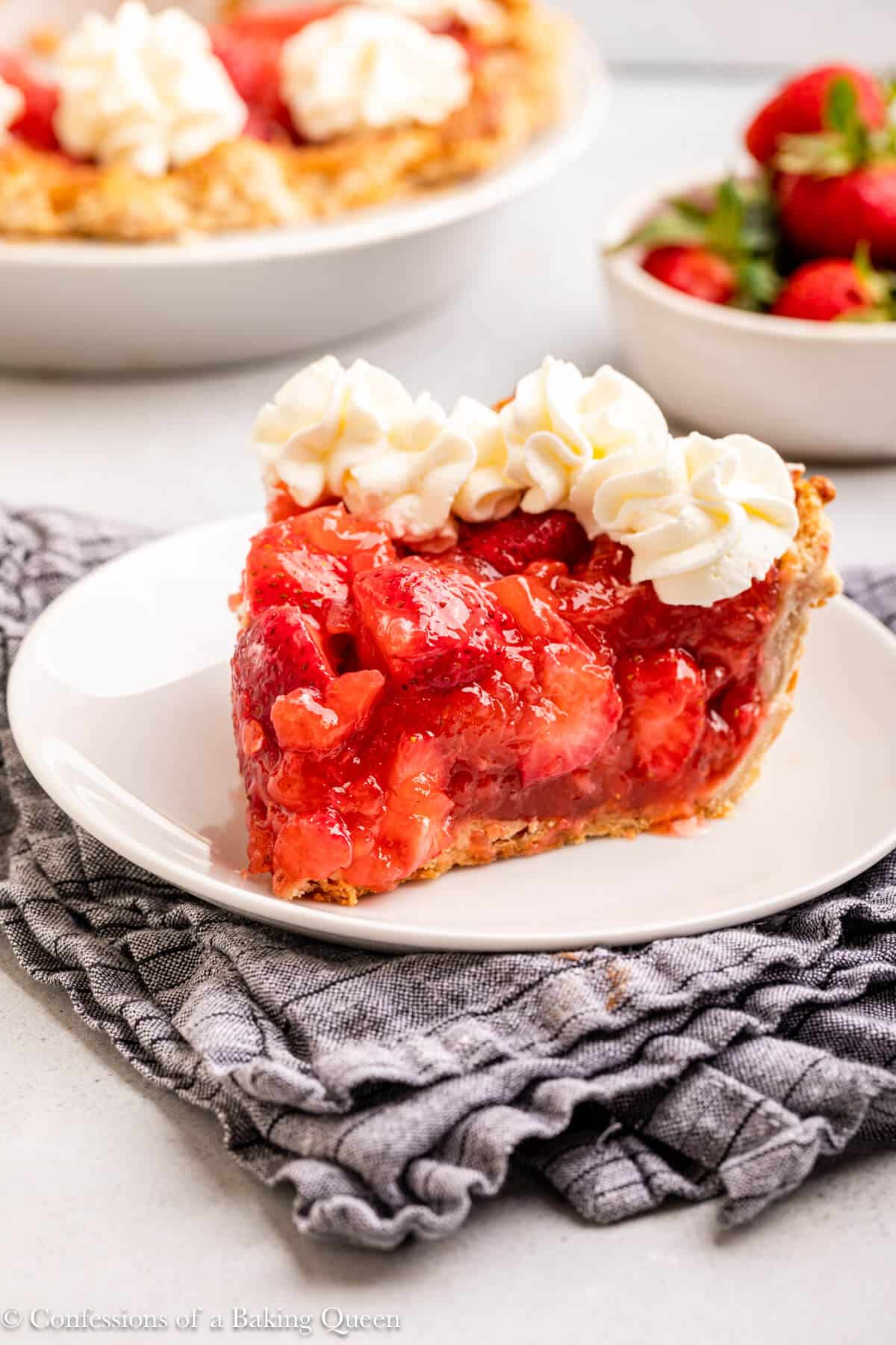 slice of strawberry pie with whipped cream on top on a white plate on a blue linen on a grey surface