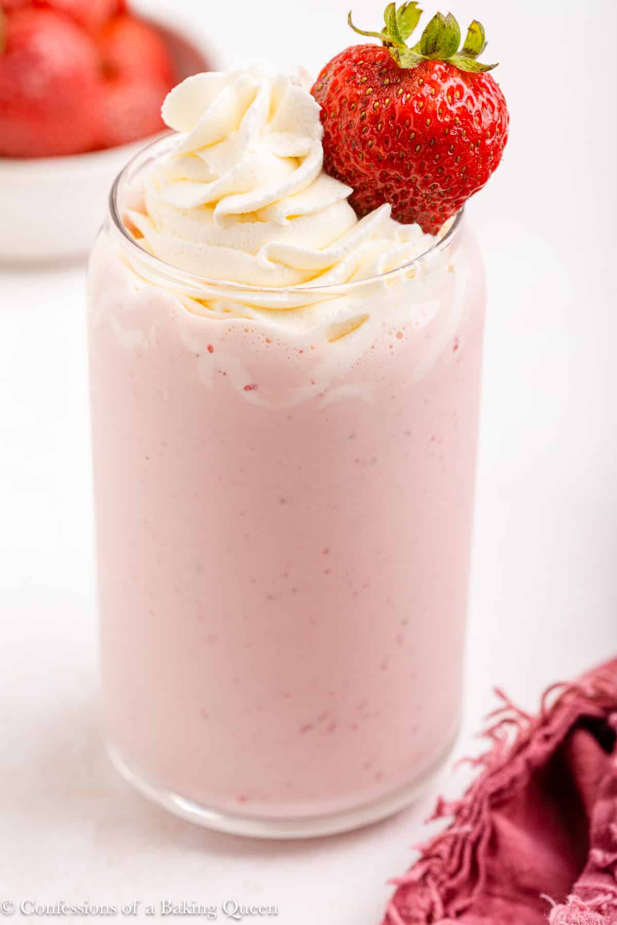 strawberry milk shake with whipped cream in a glass on a light surface with a pink linen and bowl of strawberries
