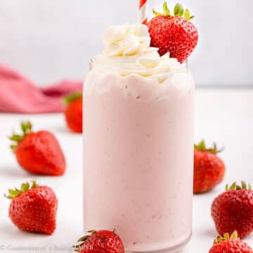strawberry milkshake with whipped cream and a berries in a tall glass with a red and white straw on a light surface.