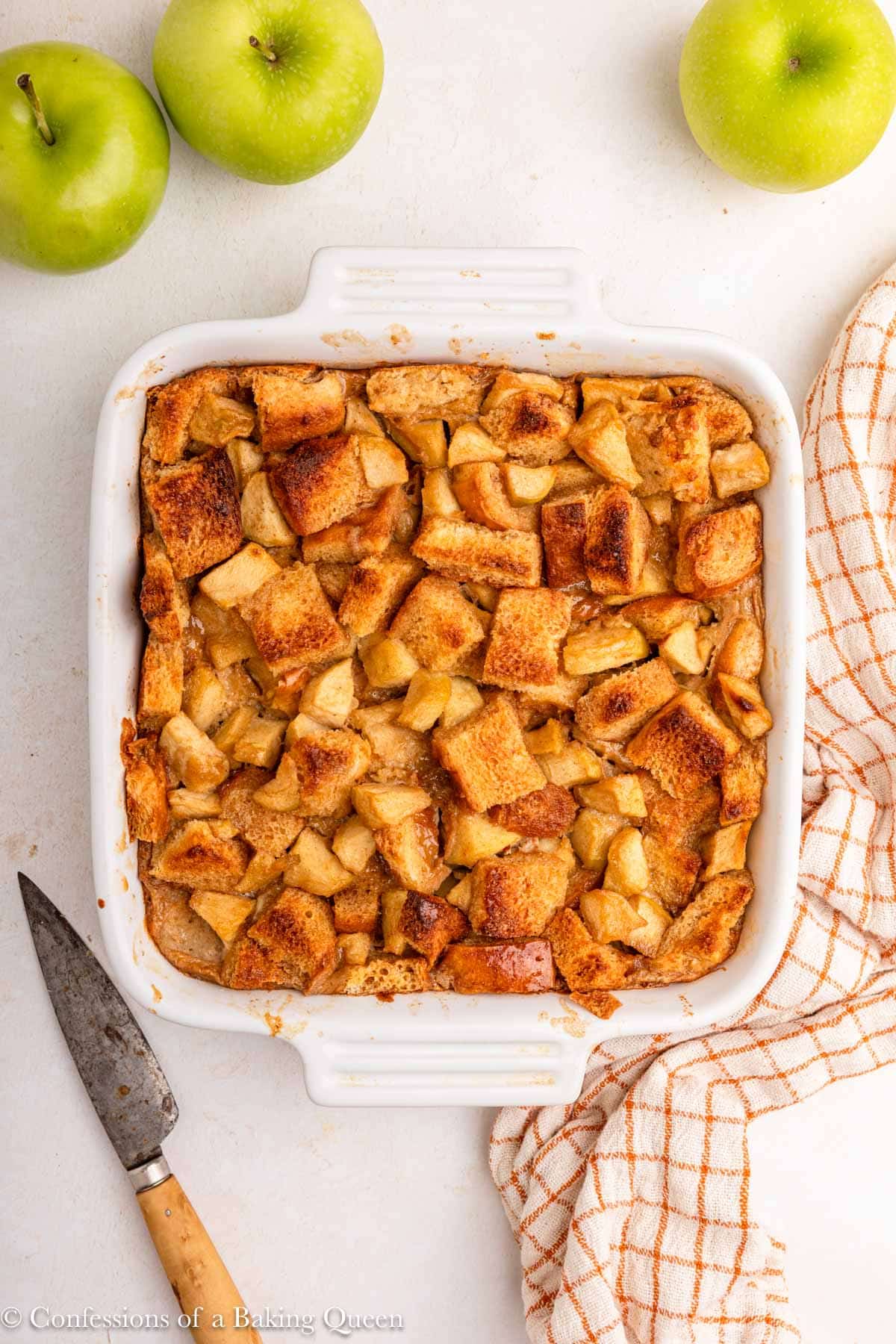 apple bread pudding just baked on a light surface with an orange linen and apples.