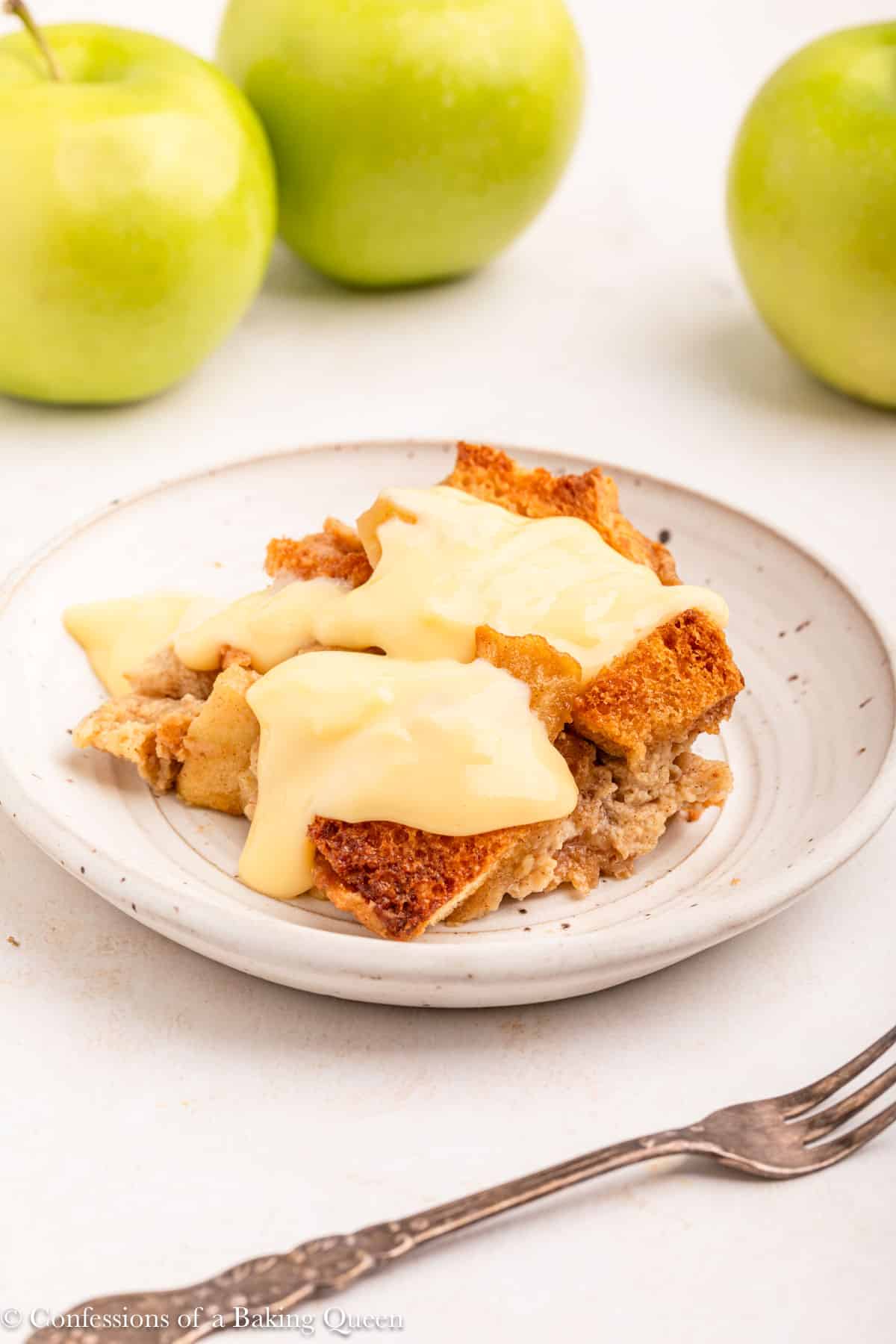 apple bread pudding slice with custard on top on a white plate on a light surface with apples