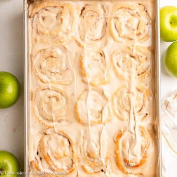 apple cinnamon rolls with maple frosting in a pan on a light surface with green apples.