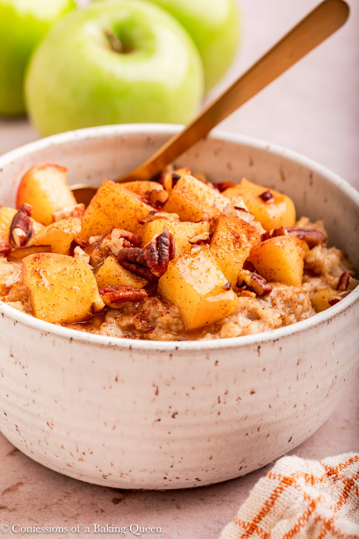 up close of apple cinnamon oatmeal with a gold spoon on a light brown surface with apples in the background.
