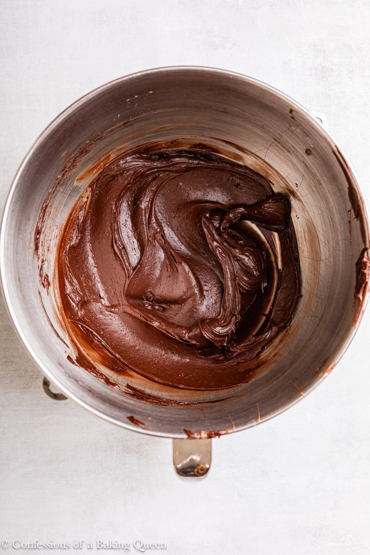 fudgy chocolate frosting in a large metal bowl on a light grey surface.