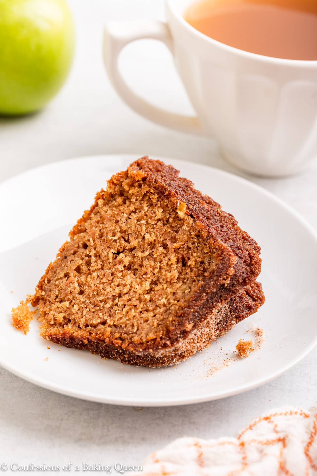slice of apple cider cake on a white plate next to a cup of apple cider, apple and orange linen on a grey surface.